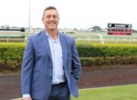 Smiling man in a blue suit standing outdoors at a racecourse with a scoreboard in the background