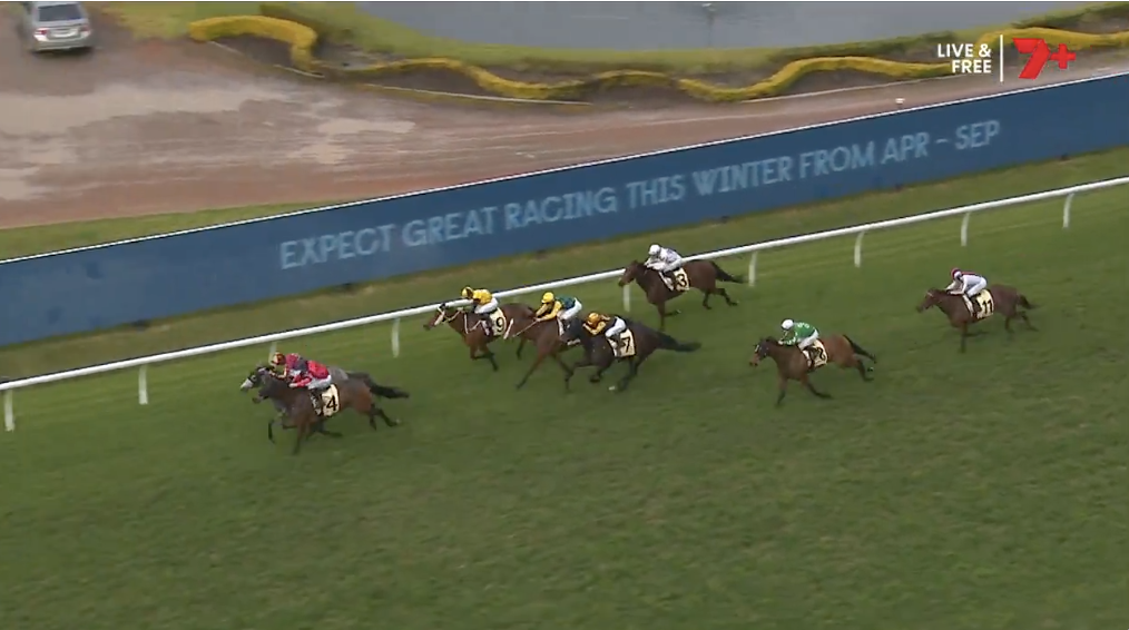 Horses and jockeys sprint along a turf racecourse near the inner rail with a blue advertising banner in the background.