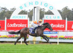 Jockey in orange and blue silks riding a dark horse over a hurdle at a Ladbrokes-sponsored race track.