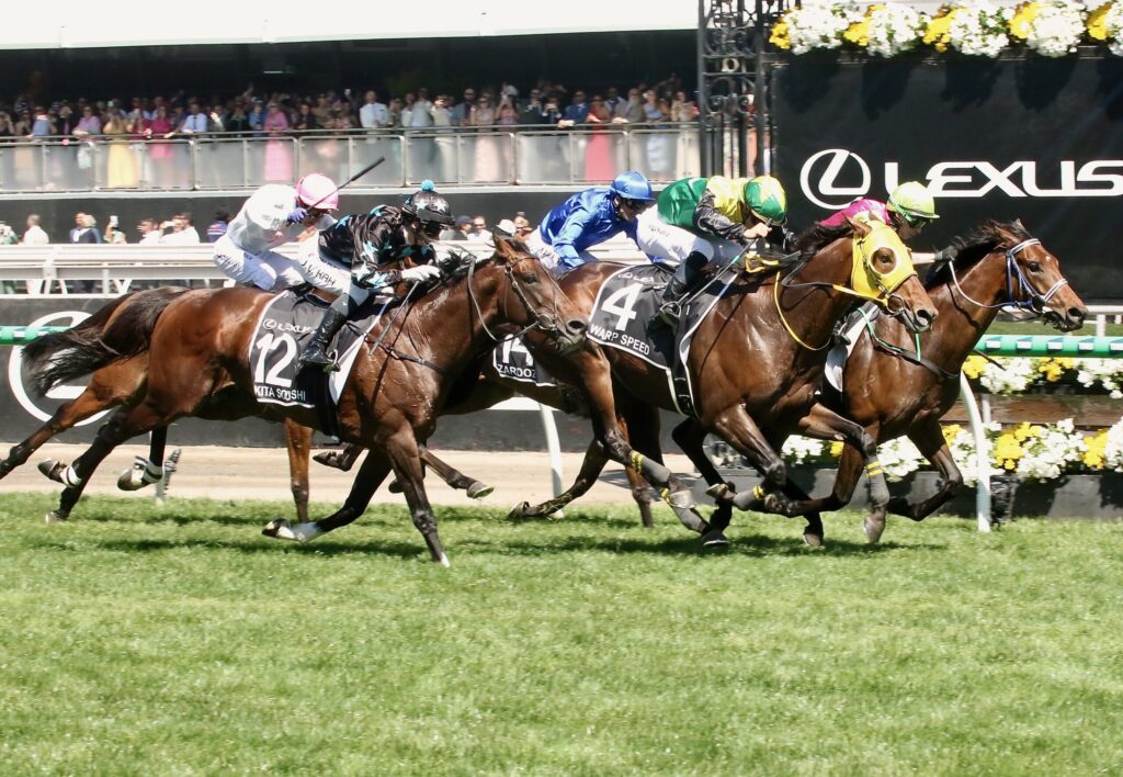 Jockeys in colorful silks race side by side on thoroughbreds with stands and spectators in the background.