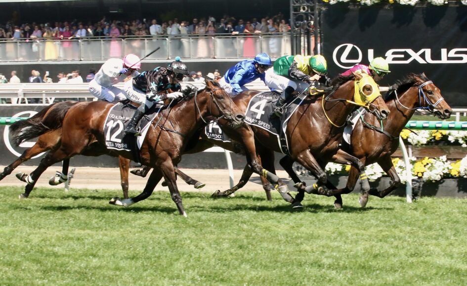 Jockeys in colorful silks race side by side on thoroughbreds with stands and spectators in the background.