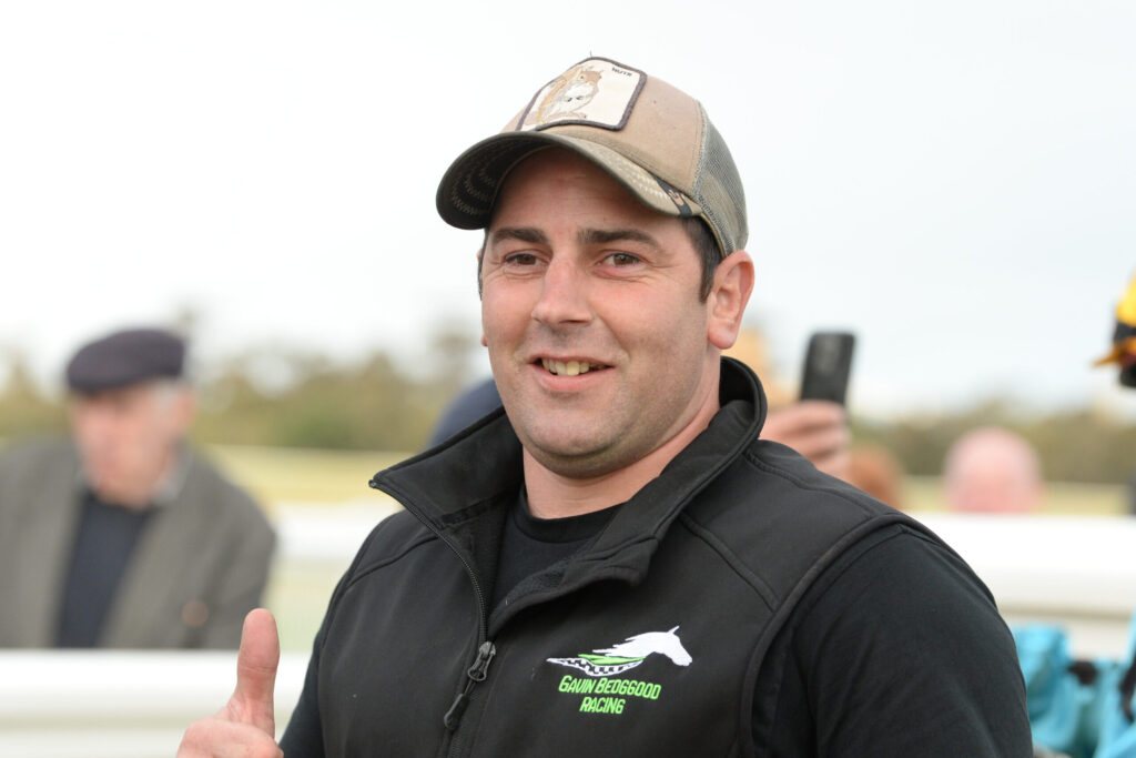Smiling man in a beige cap and black jacket giving a thumbs up at an outdoor event, with a blurred crowd in the background.