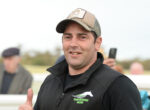 Smiling man in a beige cap and black jacket giving a thumbs up at an outdoor event, with a blurred crowd in the background.