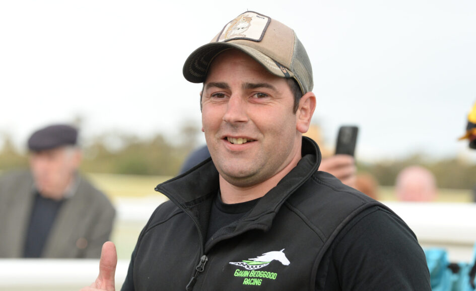 Smiling man in a beige cap and black jacket giving a thumbs up at an outdoor event, with a blurred crowd in the background.