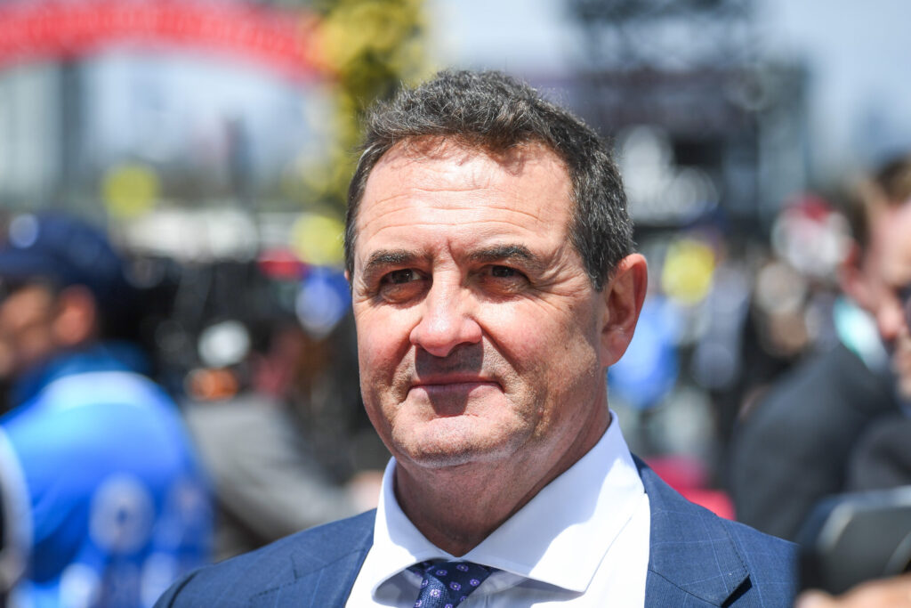 Close-up portrait of a man in a blue suit and patterned tie at an outdoor event, with a blurred crowd in the background.
