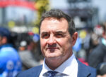 Close-up portrait of a man in a blue suit and patterned tie at an outdoor event, with a blurred crowd in the background.