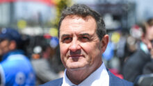 Close-up portrait of a man in a blue suit and patterned tie at an outdoor event, with a blurred crowd in the background.