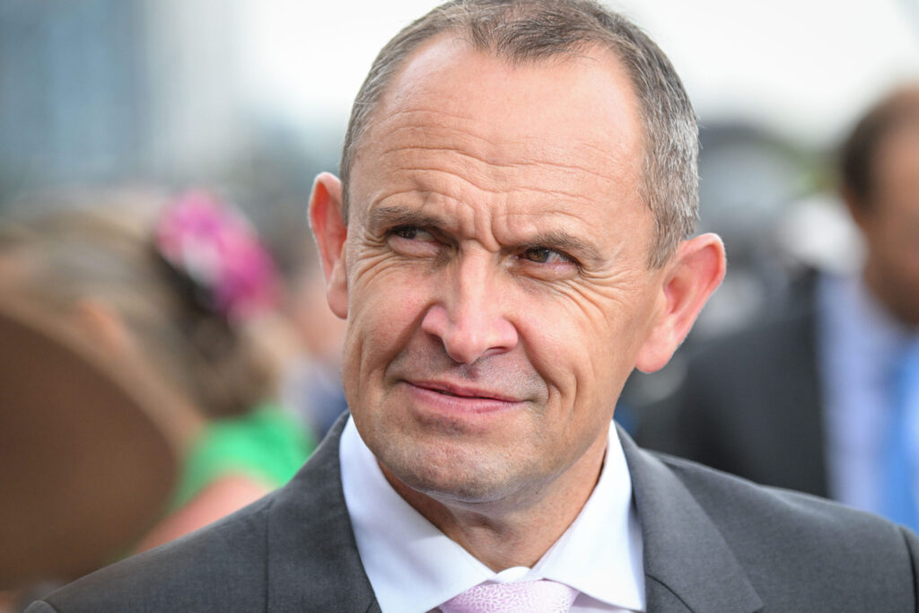 Close-up of a middle-aged man in a dark suit and pink tie at an outdoor event, smiling slightly.