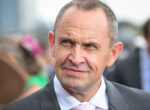 Close-up of a middle-aged man in a dark suit and pink tie at an outdoor event, smiling slightly.