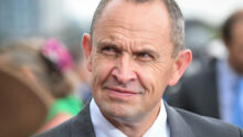 Close-up of a middle-aged man in a dark suit and pink tie at an outdoor event, smiling slightly.