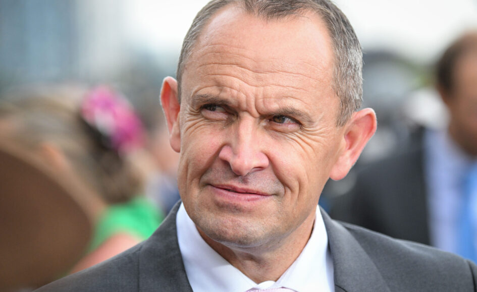 Close-up of a middle-aged man in a dark suit and pink tie at an outdoor event, smiling slightly.